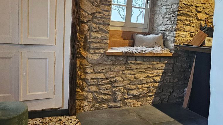 A cozy room with a stone wall and wooden ceiling. The floor is designed with colorful tiles and a window offers a view outside.