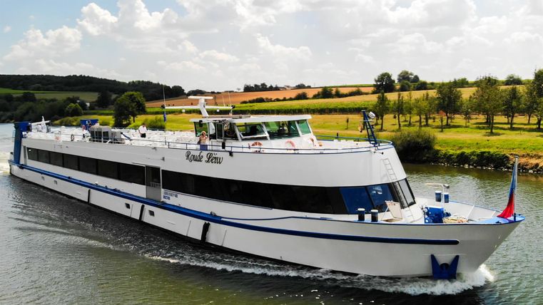 A white ship sails on a calm river. In the background, green fields and trees can be seen.