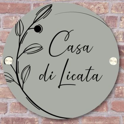 Ein runder Schild mit der Aufschrift "Casa di Licata" und einem minimalistischen Pflanzenmotiv. Der Hintergrund ist grau und der Schild hat metallische Details.