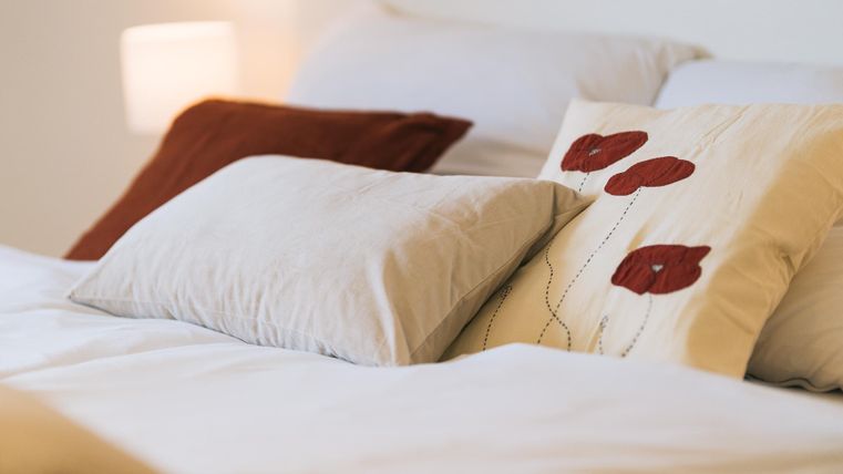 Een gezellig bed met meerdere kussens. Een kussen heeft een bloemenpatroon en het beddengoed is licht en vrolijk.