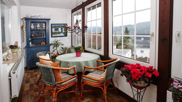 Ein gemütlicher Wintergarten mit einem runden Tisch und Stühlen. Große Fenster bieten einen Blick ins Freie und es gibt einige Pflanzen sowie dekorative Elemente.