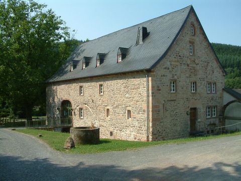 Ein charmantes, steinernes Gebäude mit einem schiefergedeckten Dach. Darum herum gibt es eine grüne Landschaft und einen runden Brunnen.