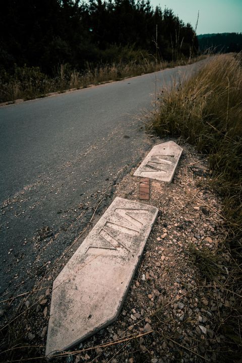 Ein verlassenes Stück Straße mit zwei beschädigten Fahrbahnmarkierungen. Umgeben von Gras und Bäumen im Hintergrund.