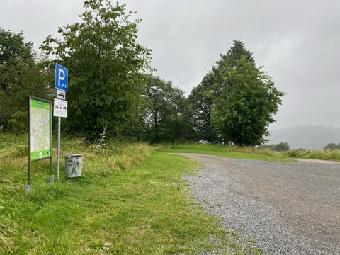 Een parkeerplaats in de natuur, omringd door bomen en gras. De lucht is bewolkt en de weg leidt het groen in.