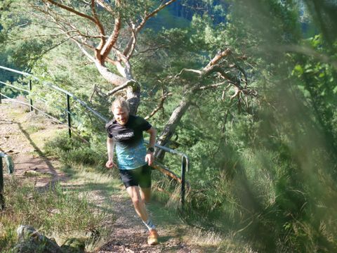 Een man jogt op een smal bospad. Op de achtergrond zijn bomen en een heldere lucht te zien.