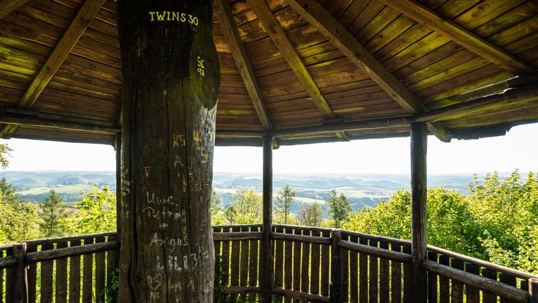 Ein Aussichtspunkt mit Holzverkleidung und einer Säule in der Mitte. Man sieht grüne Hügel und eine weite Landschaft im Hintergrund.