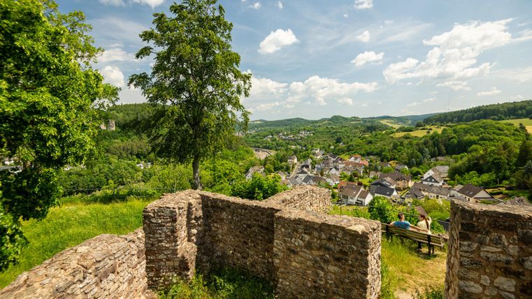 Ruinenmauer der Löwenburg mit Blick auf ein Dorf und grüne Hügel. Zwei Personen sitzen auf einer Bank. Blauer Himmel mit Wolken.