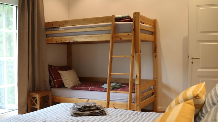A cozy bedroom with a wooden bunk bed and a large double bed. The room has large windows and an inviting atmosphere.