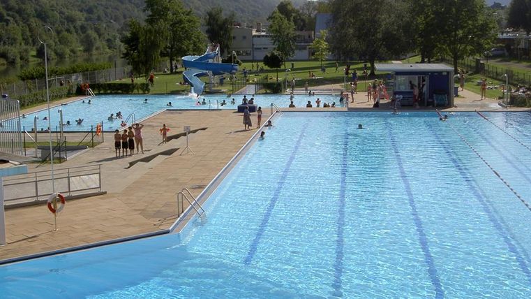 A swimming pool with clear, blue water and many guests. In the background, slides and green areas can be seen.