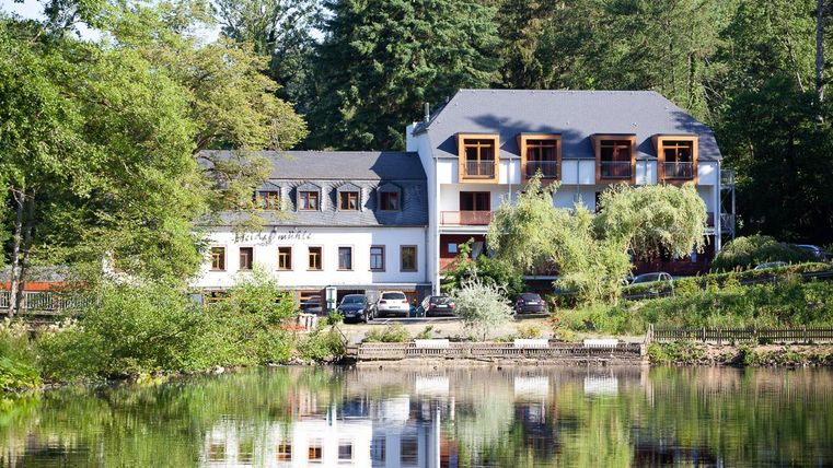 Ein malerisches Hotel am Ufer eines ruhigen Sees, umgeben von Bäumen. Die reflektierende Wasseroberfläche verleiht der Szenerie eine friedliche Atmosphäre.