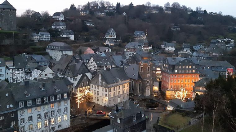 Eine malerische Stadtansicht mit traditionellen Häusern und sanften Hügeln im Hintergrund. Die Lichter der Gebäude strahlen in der Dämmerung und schaffen eine gemütliche Atmosphäre.