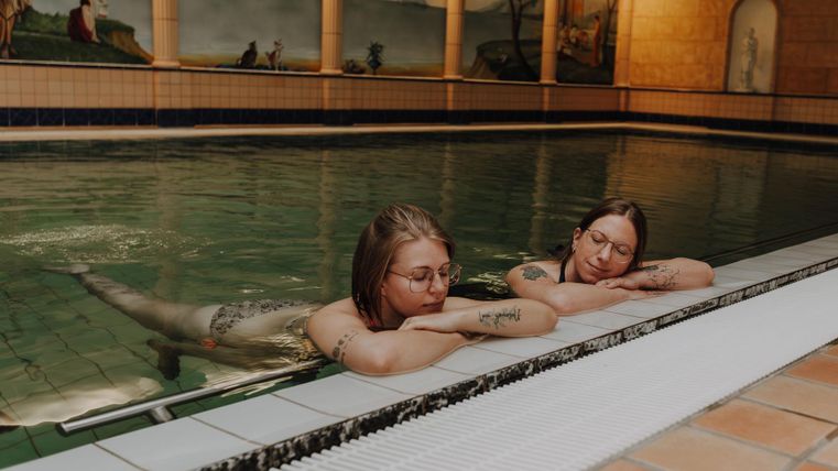 Zwei Frauen entspannen am Beckenrand in einem Schwimmbad. Das Wasser ist ruhig und die Atmosphäre ist friedlich.
