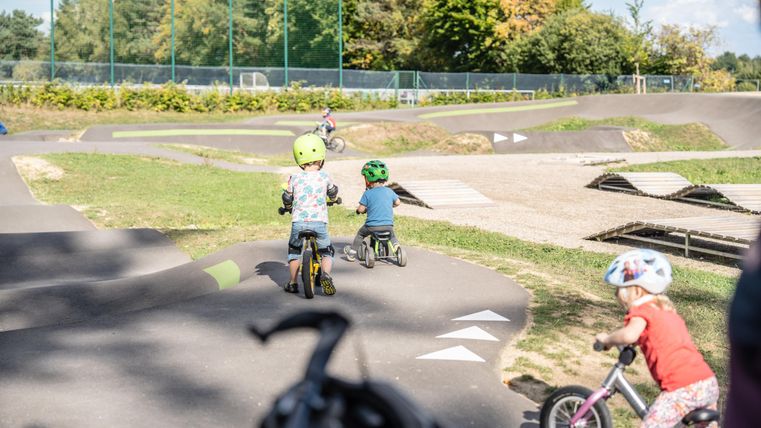 Kinder fahren auf Fahrrädern auf einem Fahrradpark. Die Umgebung ist grün und sonnig, ideal zum Spielen und Üben.