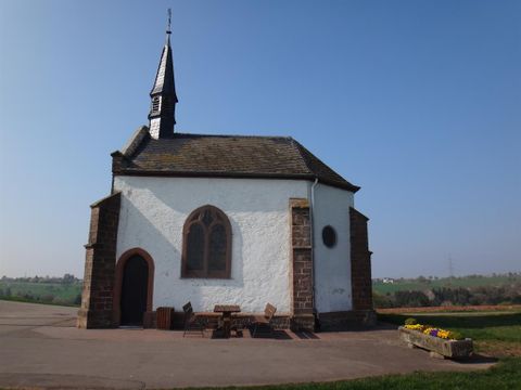 Eine kleine, charmante Kapelle mit einem spitzen Dach und einem Glockenturm. Umgeben von einer ruhigen Landschaft und bedeckt mit klarem Himmel.