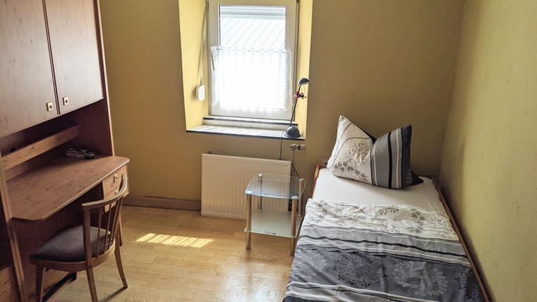 A simple room with a bed, a table, and a chair. The window lets in a lot of daylight.