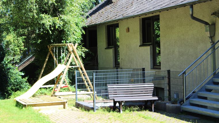Ein Spielbereich mit einer Schaukel und einer Rutsche steht neben einem Gebäude. Im Hintergrund sind üppige Bäume zu sehen.