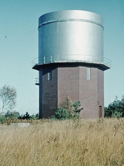 A tall water tower made of bricks with a metallic upper part. It stands in an open, green landscape with tall grass.