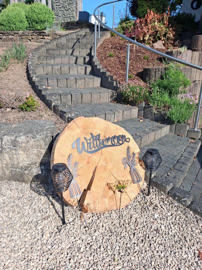 A wooden sign with the inscription "Welcome" stands by a staircase. In the background, plants and stones can be seen.
