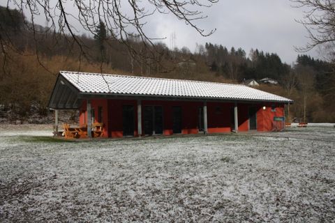 Ein rotes Gebäude mit einem weißen Dach steht in einem schneebedeckten Feld. Im Hintergrund sind Bäume und sanfte Hügel sichtbar.
