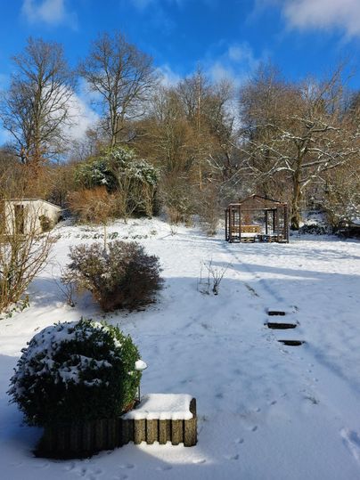 Ein verschneiter Garten mit einem Pavillon im Hintergrund. Die Bäume sind mit Schnee bedeckt und der Himmel ist blau mit einigen Wolken.