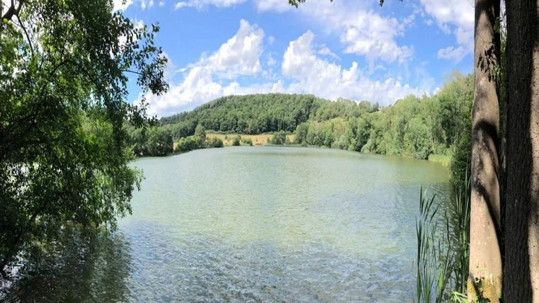 A tranquil lake surrounded by lush greenery and gentle hills. The blue sky is streaked with white clouds.
