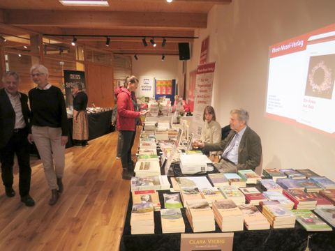 Een boekenstand met verschillende boekomslagen en meerdere mensen die zich bij de stand bevinden. Op de achtergrond zijn andere stands en een presentatie te zien.