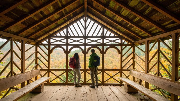 Twee wandelaars staan in een houten hut met grote ramen en bewonderen het herfstlandschap. De hut is eenvoudig, maar uitnodigend en biedt een mooi uitzicht.