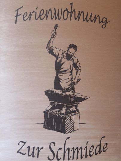 Eine Kunschrift mit der Aufschrift „Ferienwohnung Zur Schmiede“. Darauf ist eine stilisierte Darstellung eines Schmieds zu sehen, der an einemamboss arbeitet.