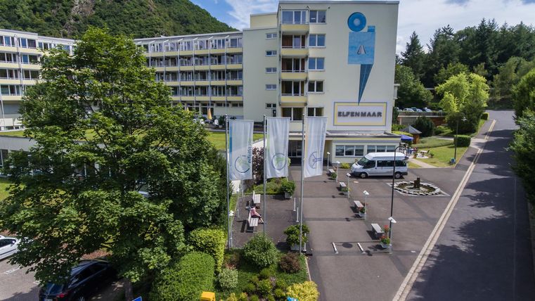 Een modern gebouw met een grote parkeerplaats en omringende bomen. De lucht is gedeeltelijk bewolkt en de omgeving ziet er uitnodigend uit.