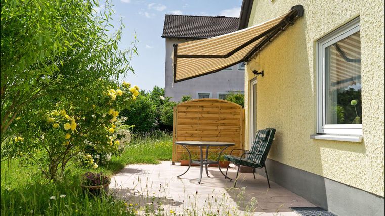 Eine gemütliche Terrasse mit einem Tisch und Stühlen. Ein Sonnenschutz ist ausgefahren, um Schatten zu spenden.