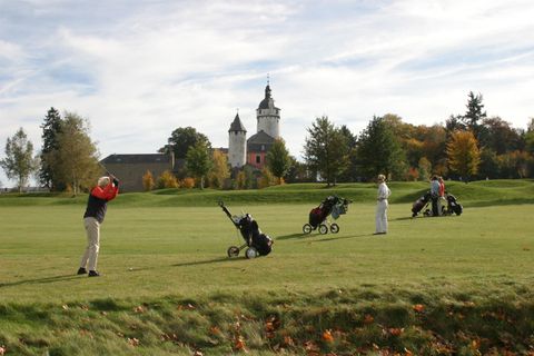 A group of golfers is playing on a beautiful golf course overlooking a historic building. The sky is clear and the surroundings are surrounded by trees and autumn leaves.