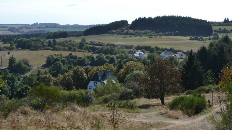 Eine ruhige Landschaft mit sanften Hügeln und Bäumen. Im Vordergrund ist ein Weg, der zu einem kleinen weißen Haus führt.