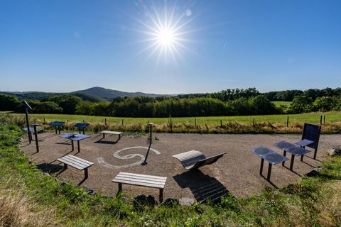 Ein Aussichtspunkt mit interaktiven Stationen und Bänken. Im Hintergrund ist eine grüne Landschaft und ein klarer blauer Himmel zu sehen.