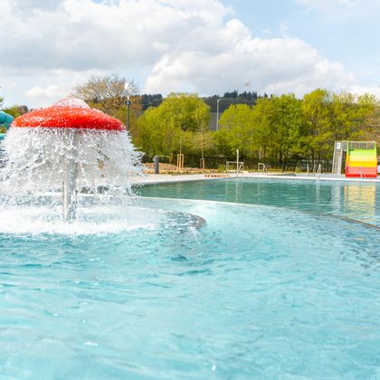 Ein Schwimmbad mit klarem Wasser und einem bunten Wasserfall. Im Hintergrund sind Bäume und ein hellblauer Himmel zu sehen.