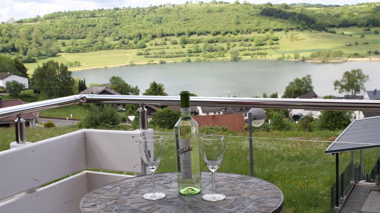 Ein schöner Ausblick von einem Balkon auf einen ruhigen See und grüne Landschaft. Auf dem Tisch stehen eine Flasche Wein und zwei Gläser.