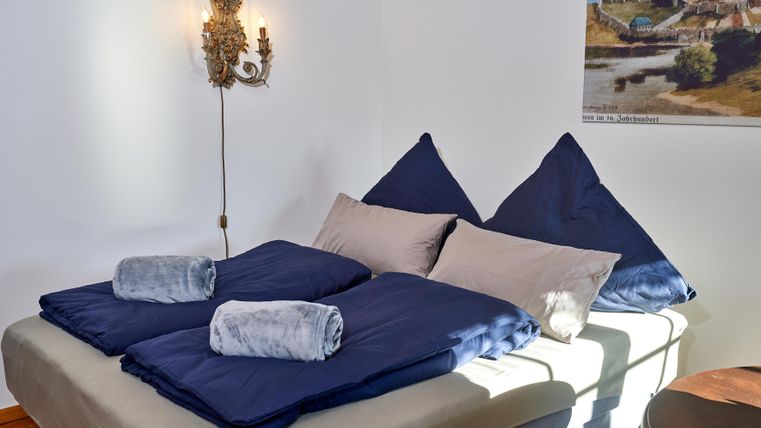 A cozy bedroom with a large bed and blue bedcovers. In the background, a mural and a decorative lamp can be seen.