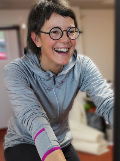 Eine fröhliche Frau beim Training auf einem Fahrrad. Sie trägt einen grauen Kapuzenpullover und lacht herzlich.
