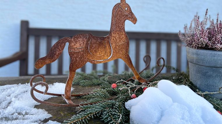 Ein dekoratives Pferd aus Holz steht auf einer geschneiten Oberfläche. Im Hintergrund sieht man eine Holzbank und eine Pflanze in einem Topf.