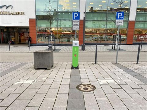 A modern parking ticket machine stands in a paved square in front of a large building. In the background, there are parking spaces and a large glass façade.