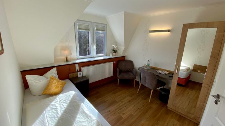 Ein helles Zimmer mit einem Einzelbett, einem Stuhl und einem Tisch. Große Fenster sorgen für viel Tageslicht und eine gemütliche Atmosphäre.