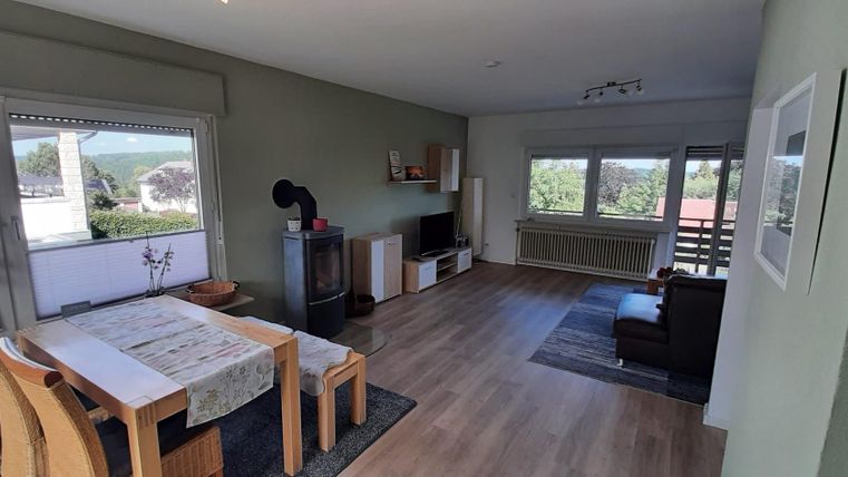 Ein helles Wohnzimmer mit einem Esstisch und Stühlen. Große Fenster bieten einen Blick auf die Natur draußen.