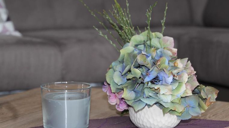 A decorative arrangement with colorful hydrangeas in a white vase is on a table. Next to it, there is a glass with a gray candle.