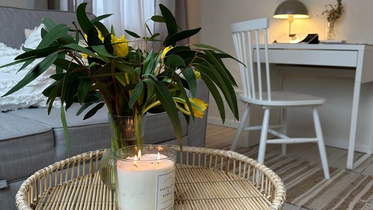 Een gezellige woonkamer met een ronde bijzettafel en een vaas met verse bloemen. Op de achtergrond staat een bureau met een lamp en lichte gordijnen.