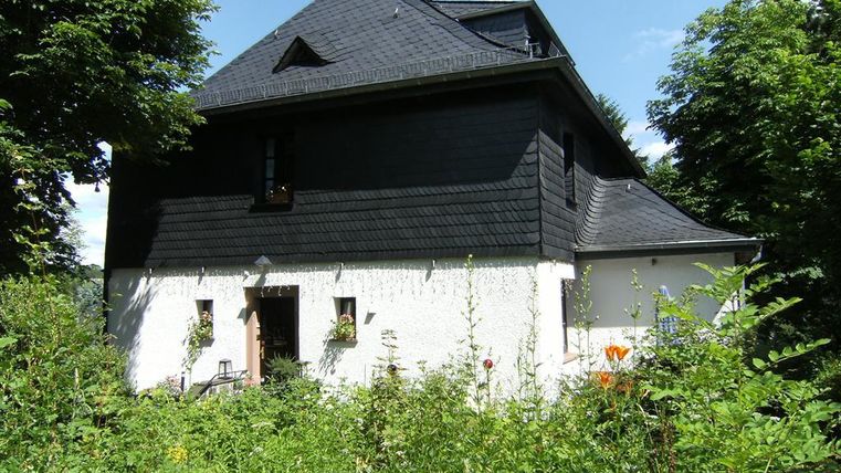 Een charmant zwart huis met een witte onderkant in een groene omgeving. Omgeven door weelderige planten en bomen straalt het een rustige sfeer uit.