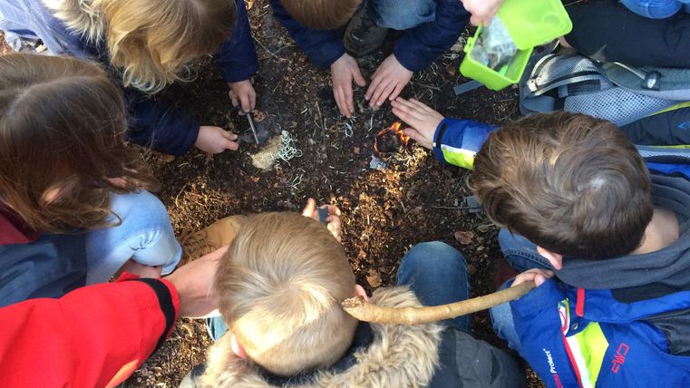 Kinder sitzen im Kreis auf Waldboden und versuchen, mit Werkzeugen ein Feuer zu entfachen. Ein Erwachsener hilft mit einer grünen Box.