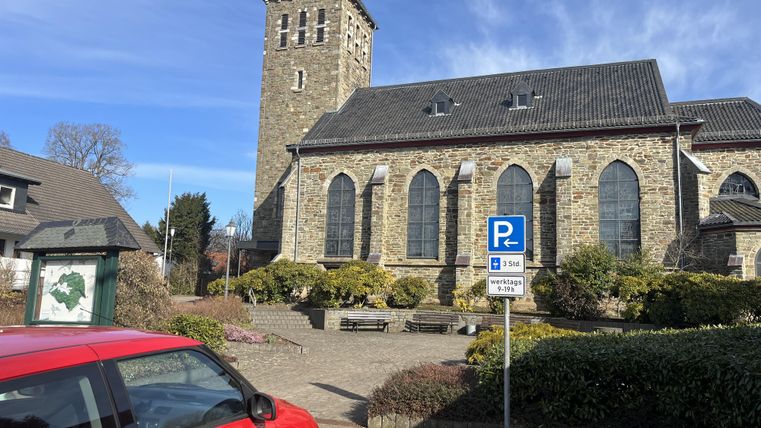 Een oude kerk met een markante toren staat op de voorgrond. Op de voorgrond staat een rode auto en rondom zijn verzorgde planten en een parkeerzone te zien.