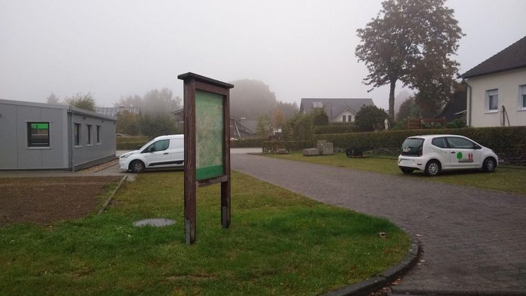 Ein nebliger Ort mit einer Informationstafel und zwei Autos auf dem Parkplatz. Im Hintergrund sind einige Gebäude und Bäume sichtbar.