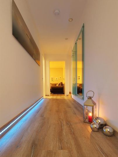 Een lichte gang met houten vloeren en indirecte verlichting. Aan het einde van de gang is een gezellige woonkamer zichtbaar, en een lantaarn met een kaars zorgt voor een uitnodigende sfeer.