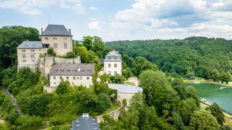 Ein historisches Schloss umgeben von grünen Wäldern und einem klaren See. Der Himmel ist teilweise bewölkt.