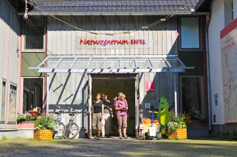 Ein Eingangsbereich des Naturzentrums Eifel mit zwei Personen, die draußen stehen. Das Gebäude hat eine moderne Holzfassade und ist von Pflanzen umgeben.